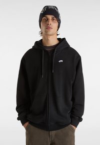 Czarna bluza z kapturem na zamek błyskawiczny, z kieszenią kangurką, kapturem ze sznurkiem i małym białym logo na piersi, noszona z wzorzystą czapką beanie w czarnym kolorze.