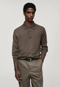 Polo shirt en tricot marron à manches longues avec un col classique et une patte de boutonnage à trois boutons, associé à un pantalon beige clair et une ceinture noire.