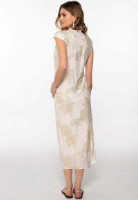 Robe longue sans manches de couleur beige avec de subtils motifs floraux dorés et une surface texturée. Fente sur le côté et coupe décontractée. Sandales à lanières aux pieds.