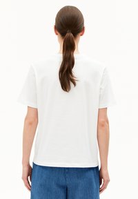 Femme avec de longs cheveux bruns attachés en queue de cheval, portant un t-shirt blanc à manches courtes uni et un jean bleu, se tenant de dos.