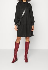 Modellen står i en svart lager-på-lager-klänning och burgundy knähöga stövlar, med en liten svart crossbody-väska. Den neutrala bakgrunden framhäver outfiten.