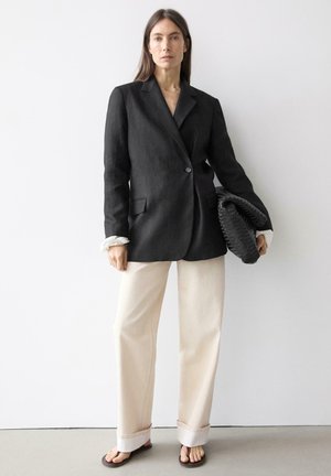 Žena stojící u bílé zdi, oblečená v černém saku, béžových kalhotách s širokými nohavicemi a žabkách, držící velké černé texturované psaníčko.