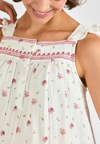 Top in tessuto leggero e cremoso con ricami floreali rosa, spalline arricciate e chiusura a bottone sullo scollo. Texture morbida e design plissettato.