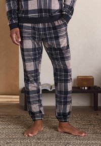 Mann, der schwarze und beige karierte Schlafanzughosen trägt, steht barfuß auf einem gewebten Teppich in einer gemütlichen Innenumgebung.