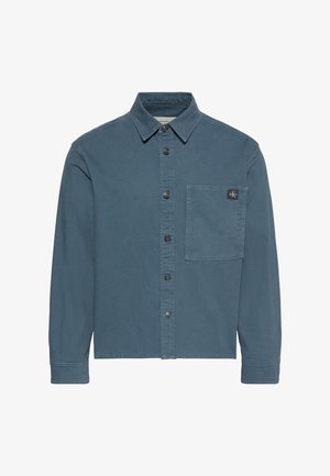 Camicia blu-grigia con bottoni, maniche lunghe, colletto e una tasca sul petto con una piccola toppa con logo nero.
