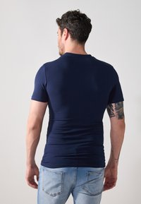 Mørkeblå tettsittende T-skjorte med korte ermer, laget av glatt stoff. Bak er det en rund hals og en subtil logo-detall nær halslinningen.