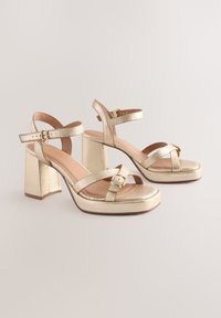 Pair of gold metallic high-heeled sandals with ankle straps, buckles, block heels, and platform soles on a light background.