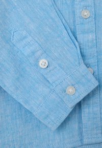 Camisa de color azul claro con una tela texturizada, puños abotonados y botones de plástico transparentes. Costuras visibles y detalle en el cuello.