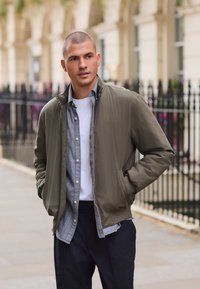 Olivgrüne Bomberjacke mit einem Reißverschluss am Kragen, darüber ein graues, gestreiftes Hemd mit Knöpfen, kombiniert mit dunklen Hosen. Außenszene.