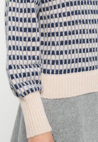 Strickpullover mit einem karierten Muster in Marineblau und Hellblau, mit einem weichen, beigen gerippten Saum und Bündchen. Das Material wirkt warm und strukturiert.