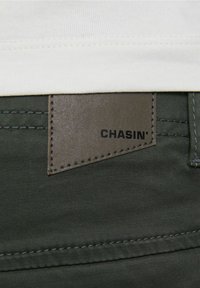 Olivově zelené kalhoty s detailem v podobě obdélníkové syntetické kožené nášivky s nápisem "CHASIN'" vyraženým černou barvou, přišité na pásku.