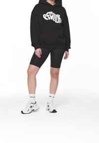 Felpa nera con logo bianco, pantaloni corti da ciclismo neri e sneakers bianche. Il modello indossa calzini bianchi con accenti neri.