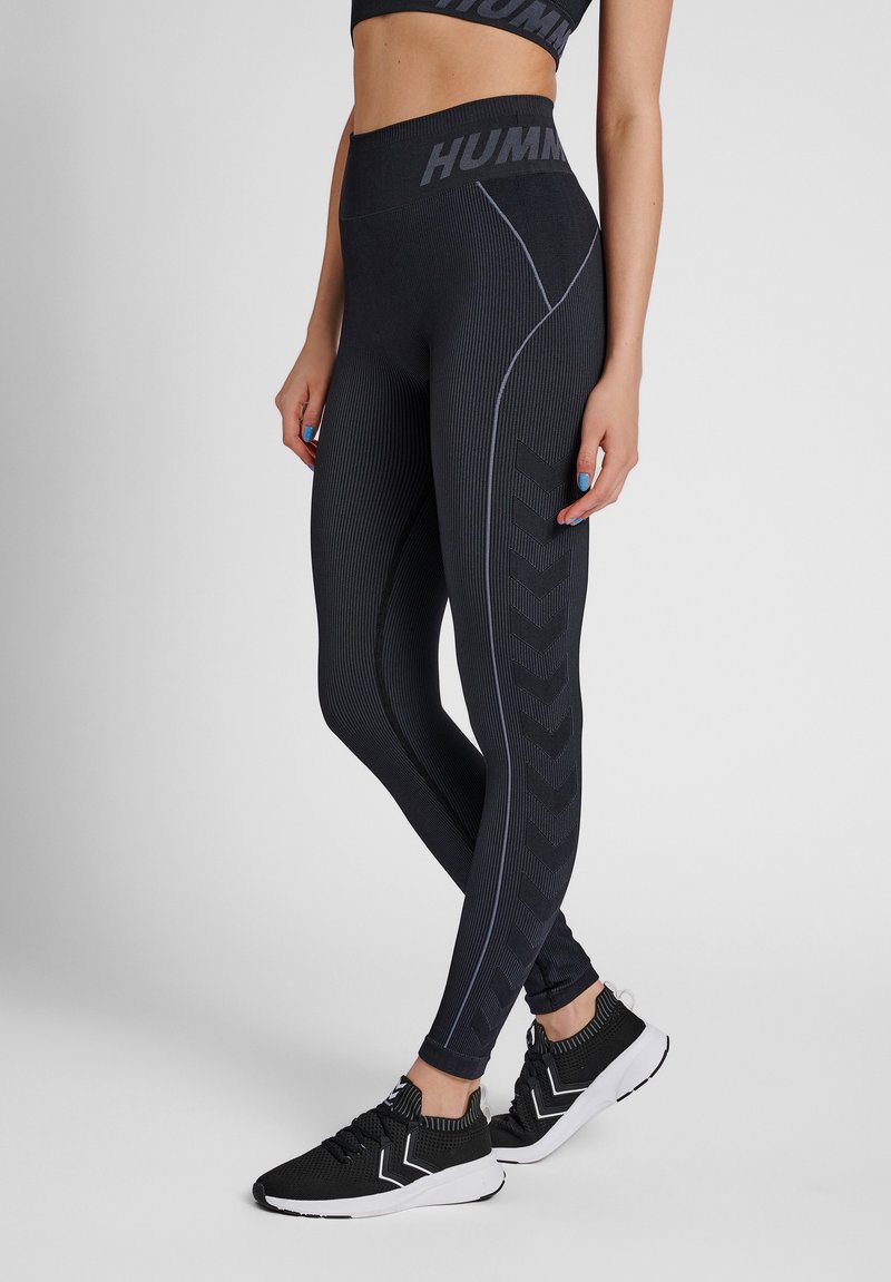 Vrouw draagt zwarte, hoog taillerengetextureerde legging met zijwaartse chevron-patronen en zwarte sportschoenen met witte zolen.