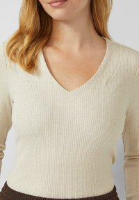 Cremefarbenes, geripptes Langarmshirt mit V-Ausschnitt. Es verfügt über eine vertikale Textur und eine figurbetonte Silhouette, kombiniert mit einem braunen Taillenbund darunter.