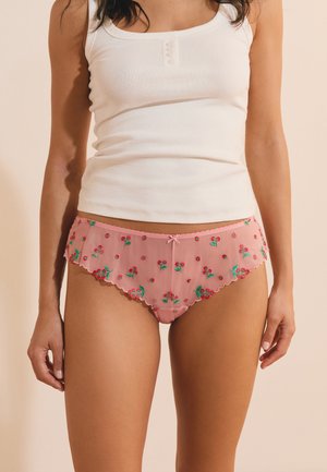 Vrouw draagt een wit mouwloos topje en roze semi-transparante slipjes met bloemenborduursel, geschulpte randen en een klein strikje vooraan in het midden.