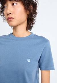 Ungdom med kort, krøllete hår som har på seg en enkel blå t-skjorte med en liten lyseblå brodert logo på brystet.