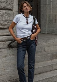 Junge Frau mit kurzen Haaren, die ein weißes T-Shirt und dunkle Jeans trägt, steht auf steinernen Stufen und lächelt mit Sonnenbrille, die am Ausschnitt hängt.