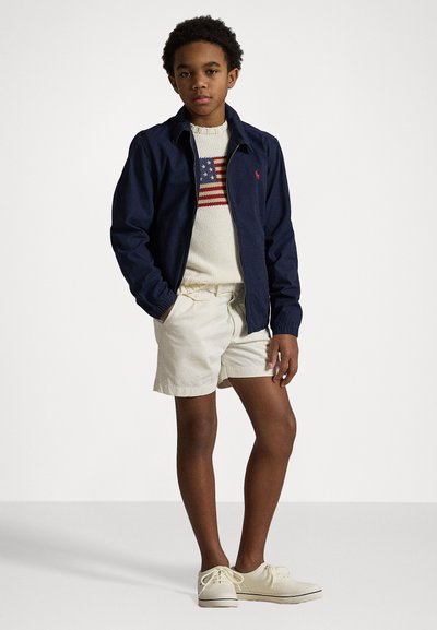 Jachetă marină peste un pulover crem cu designul steagului american, combinată cu pantaloni scurți bej și încălțăminte deschisă. Material fin cu un croi lejer.
