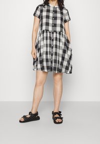 Robe à manches courtes à carreaux noir et blanc avec un devant boutonné, une jupe froncée et des poches latérales, assortie à des sandales noires à multiples lanières.