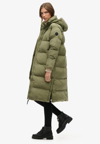 Abrigo largo de plumas en verde oliva con capucha, que cuenta con cierre de cremallera y botón a presión, bolsillos laterales y puños acanalados. Se lleva con leggins negros y botas.