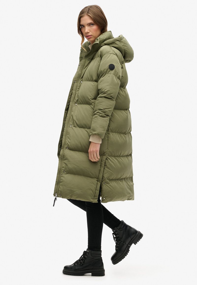 Olivgrüner langer Puffer-Mantel mit Kapuze, ausgestattet mit einem Reißverschluss und Druckknopfverschluss, seitlichen Taschen und gerippten Bündchen. Getragen mit schwarzen Leggings und Stiefeln.