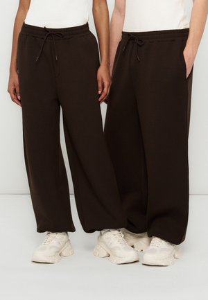 Dvaja ľudia majú na sebe tmavohnedé široké nohavice so sťahovacou šnúrkou a biele tenisky, stoja vedľa seba na bielom pozadí.