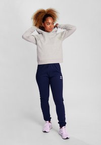 Felpa grigia con cappuccio e accenti chiari, abbinata a pantaloni jogger blu navy. L'outfit presenta polsini elastici e sneakers rosa.