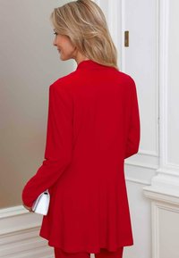 Rode blazer met een vloeiende ontwerpn en lange mouwen, voorzien van een rugplichaat. De stof lijkt glad, en het wordt gedragen met een zilveren clutch.