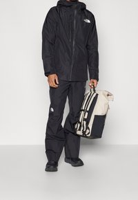 Giacca e pantaloni impermeabili neri con logo. Zaino beige con accenti e cinghie nere, progettato per l'uso all'aperto.
