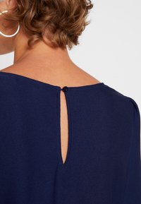 Navyblå plagg med en mjuk textur, med en baknyckelhålsöppning som säkras med en liten knapp. Minimalistisk design med rena linjer.