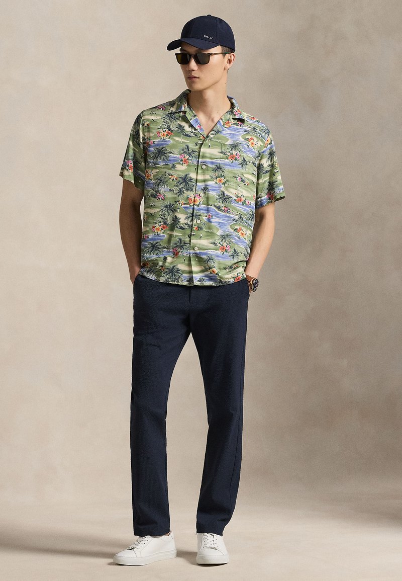 Short-sleeve floral print shirt in green with pink and blue accents, paired with dark navy trousers and white sneakers. Casual cap and sunglasses.