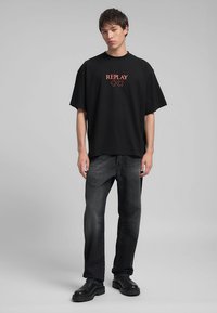 T-shirt nera oversize con logo rosso "REPLAY", abbinata a jeans dritti neri scoloriti e stivali neri con suola chunky.