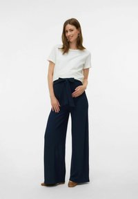 Weißes Kurzarm-T-Shirt kombiniert mit dunkelblauen weit geschnittenen Hosen mit einer vorderen Bindung. Die Schuhe sind hellbraun. Glatte Texturen überall.
