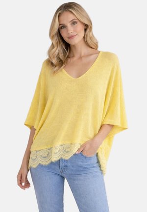 Femme portant un haut jaune ample à col en V avec bordure en dentelle et un jean bleu clair, debout, une main dans la poche, sur un fond uni.