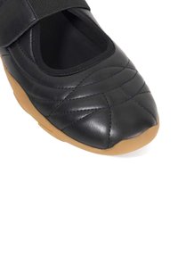 Black soft leather shoe with a quilted design and elastic strap, featuring a rounded toe and a contrasting rubber sole.