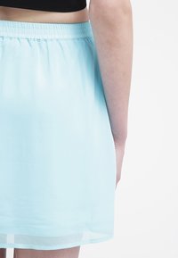Primo piano della parte bassa della schiena e del braccio di una persona che indossa una gonna azzurro chiaro e un crop top nero su uno sfondo bianco.