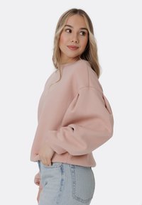 Sudadera rosa claro con cuello redondo y mangas holgadas, combinada con jeans azul claro. La tela parece suave y cómoda.