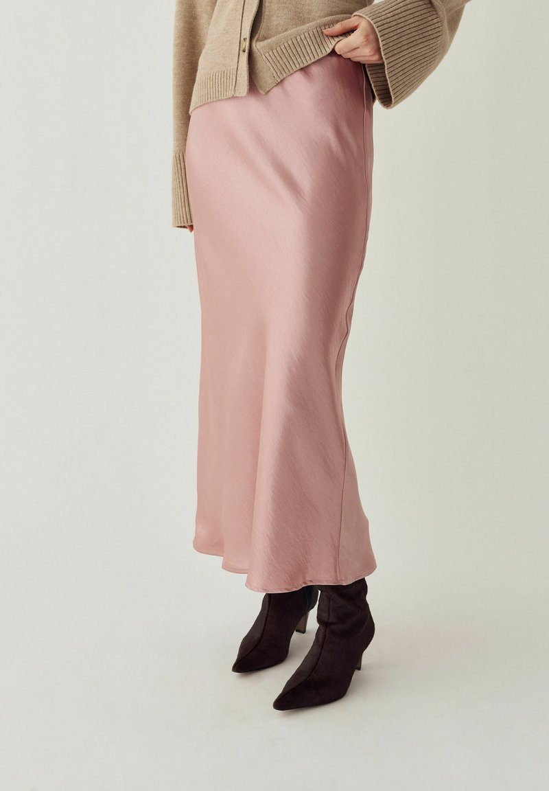 Kvinde iført beige cardigan, pink satinmidi-nederdel og sorte spidse støvler med hæl, stående mod en neutral baggrund.