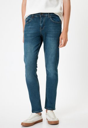 Vaqueros de mezclilla azul oscuro con corte ajustado, que cuentan con cinco bolsillos, un ligero desgastado y costuras naranjas en contraste. Combinados con zapatillas blancas.