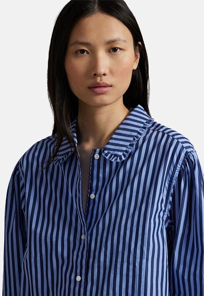 Femme aux cheveux noirs raides portant une chemise à rayures verticales bleu et marine avec un petit col à volants.