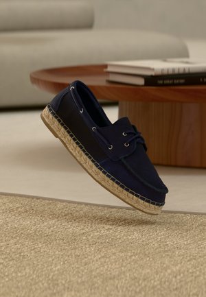 Mocassin bleu marine avec semelle en jute tressée flottant au-dessus d'un tapis beige, table basse ronde en bois et piles de livres en arrière-plan.