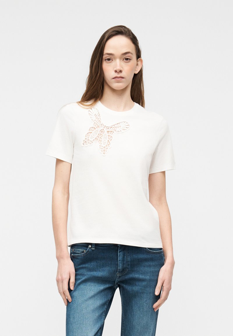 Jeune femme aux longs cheveux bruns portant un haut blanc à manches courtes avec un motif papillon en dentelle, associé à un jean en denim bleu.