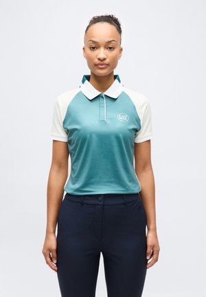 Žena s vlasmi zviazanými dozadu, oblečená v polo tričku s krátkymi rukávmi v tyrkysovej a bielej farbe s logom EA7, a tmavomodré nohavice, stojaca pred jednoliatym pozadím.