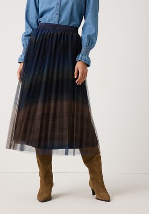 Femme portant une jupe mi-longue plissée en tulle noir, un chemisier bleu avec des poignets smockés et des bottines à talons en daim beige sur un fond uni.