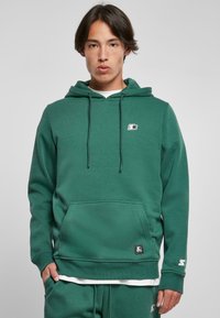 Hoodie verde in cotone con tasca a marsupio, cappuccio regolabile con cordino e patch con logo sul petto. Texture morbida, design casual.