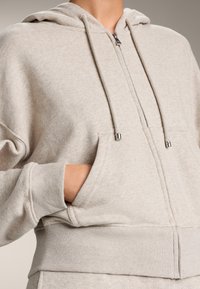 Felpa leggera grigia con zip realizzata in un tessuto morbido. Presenta una tasca frontale, polsini a coste e cappuccio regolabile con terminali metallici.