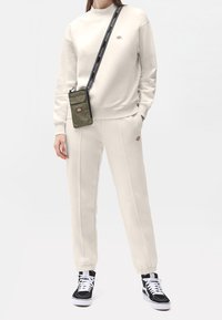 Conjunto de sudadera y pantalones de color beige claro con puños acanalados, que presenta un logotipo bordado en el pecho y en los pantalones. Combinado con zapatillas negras.