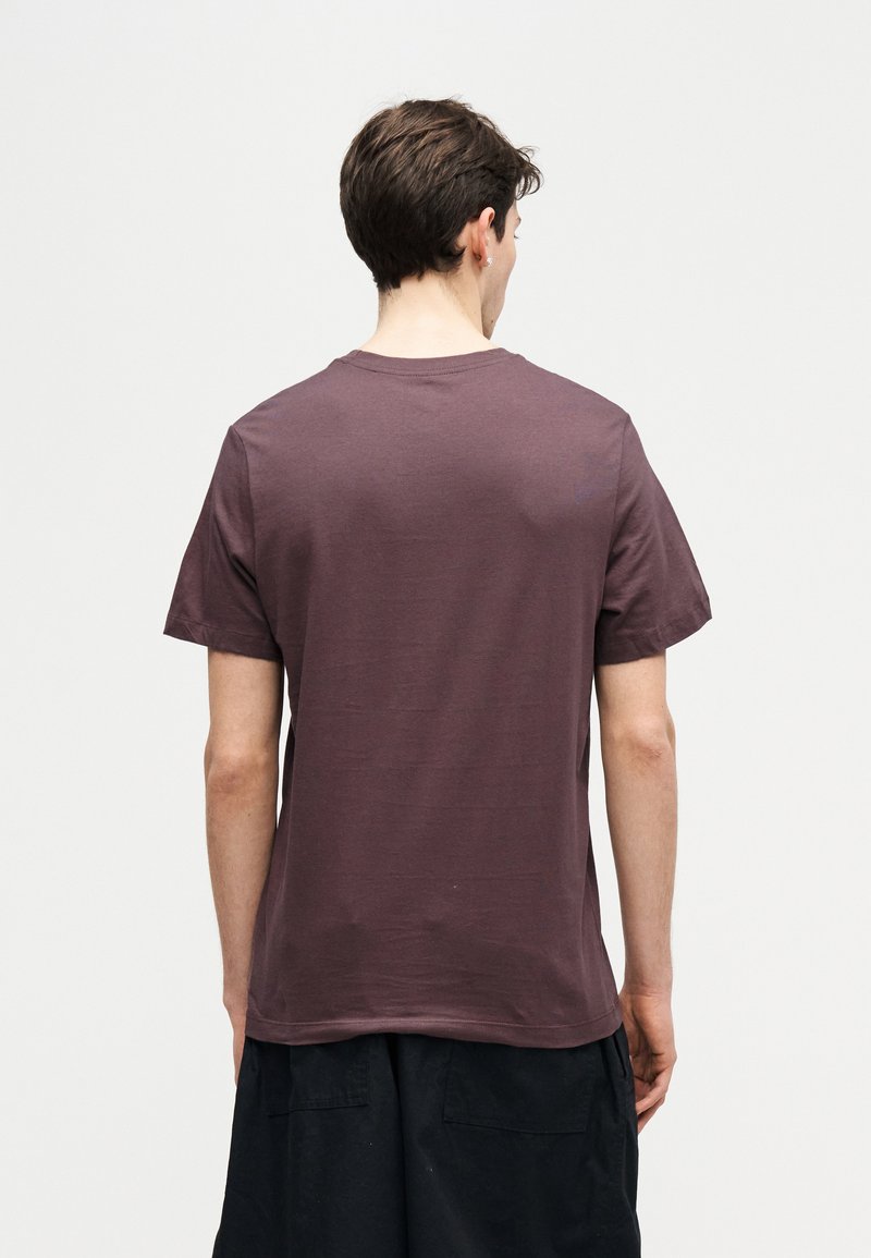 Jonge man gezien van achteren, gekleed in een effen mauve T-shirt met korte mouwen en een zwarte broek, tegen een effen achtergrond.