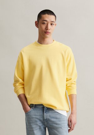Gelbes Sweatshirt mit gerippten Bündchen und Kragen, versehen mit einem dezenten, geprägten Logo auf der Vorderseite. Kombiniert mit hellblauen Jeans.
