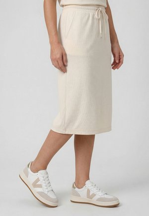 Kobieta ubrana w beżową spódnicę do kolan ze ściągaczem w talii oraz białe trampki z beżowymi i złotymi akcentami, stojąca na jasnoszarej podłodze.